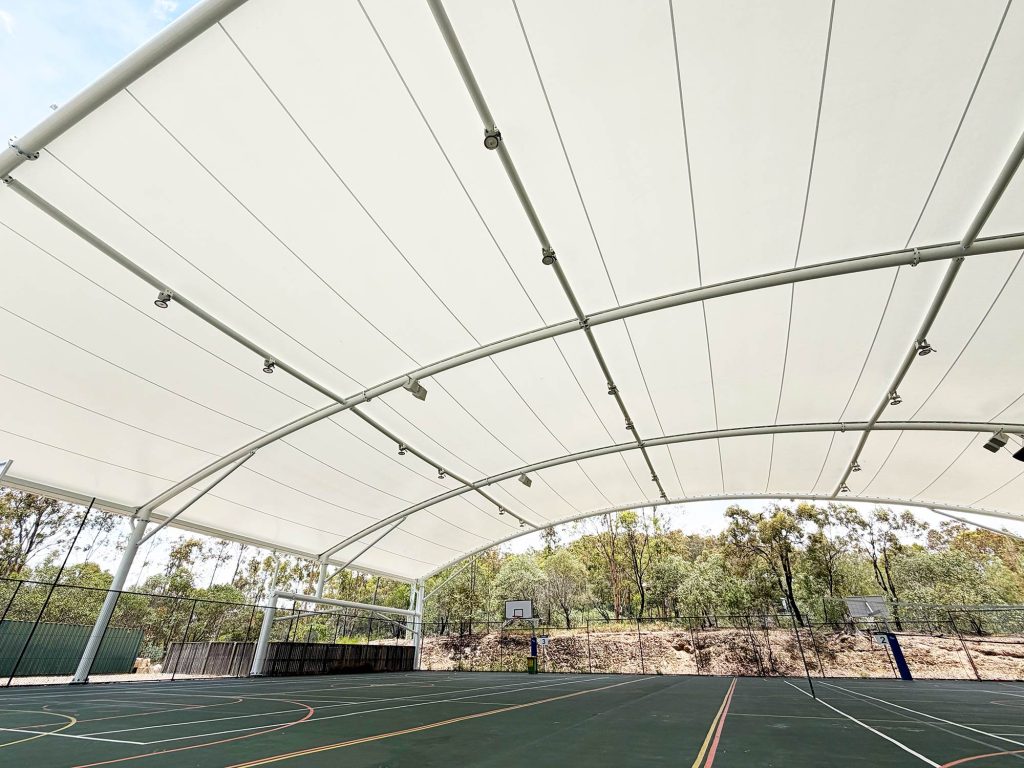 Upward-looking interior view of the white tensile shade canopy at LORDS Lutheran Ormeau Rivers District School, showing the fabric panels, cable tensioning system and floodlights against the arched steel frame, with court markings and bushland visible beyond