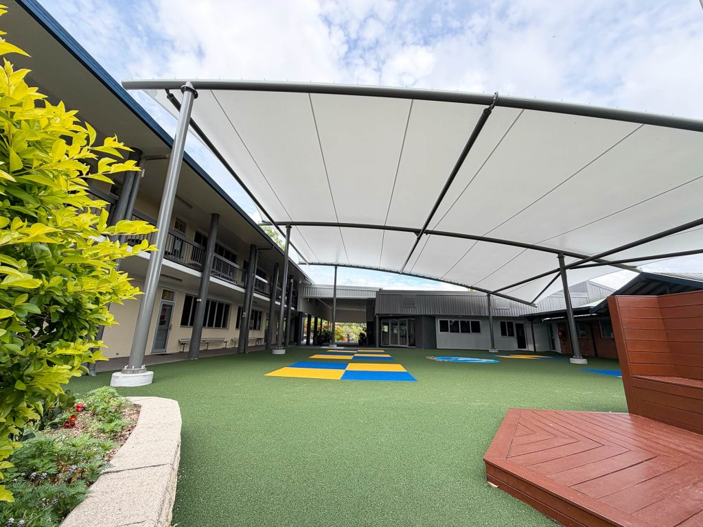 Large tensile shade canopy on a black steel frame covering a school courtyard at St Augustine's College, with artificial turf, colourful play mats, garden beds and school buildings visible beneath and around the structure