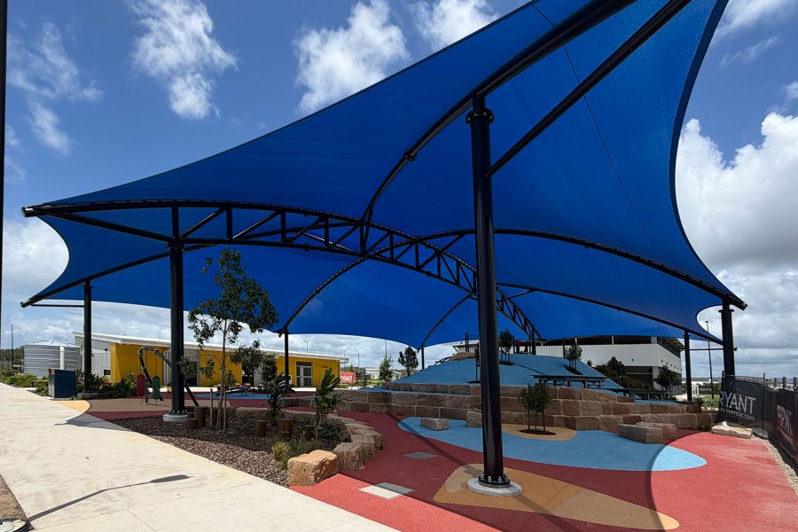Close-up ground-level view of blue hyperbolic paraboloid shade sails on black steel poles at Notre Dame College Sunshine Coast, with playground and yellow school buildings visible below