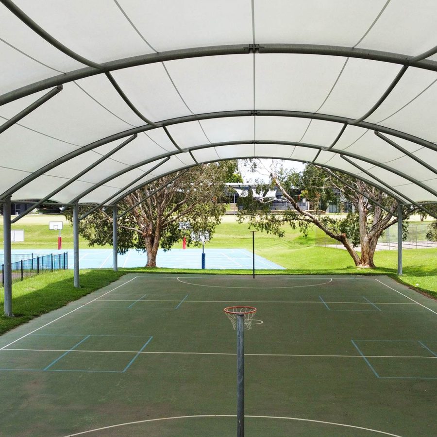 Interior end view under a large curved tensile shade canopy at Guardian Angels College, with the arched steel frame structure visible overhead, a netball and basketball court below, and lush green lawns, trees and a swimming pool visible beyond the open end of the canopy