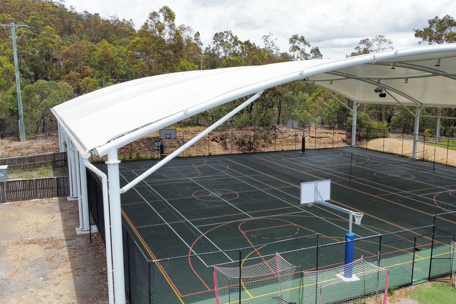 LORDS Lutheran Ormeau Rivers District School – Tensile Canopy Over Multi-Sport Court Ground-level view of the large white tensile shade canopy with floodlights covering the multi-sport basketball court at LORDS Lutheran Ormeau Rivers District School, with dense bushland in the background and court markings visible below