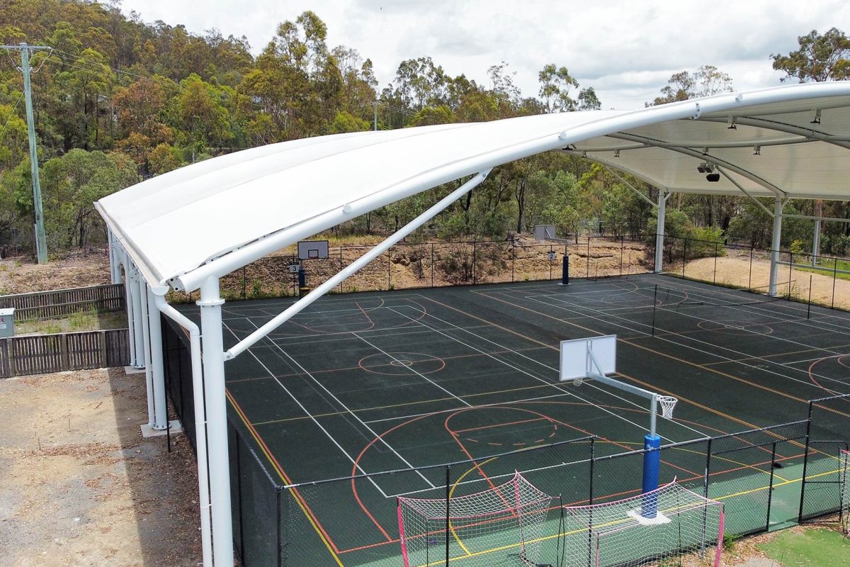 LORDS Lutheran Ormeau Rivers District School – Tensile Canopy Over Multi-Sport Court Ground-level view of the large white tensile shade canopy with floodlights covering the multi-sport basketball court at LORDS Lutheran Ormeau Rivers District School, with dense bushland in the background and court markings visible below