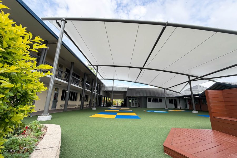 Large tensile shade canopy on a black steel frame covering a school courtyard at St Augustine's College, with artificial turf, colourful play mats, garden beds and school buildings visible beneath and around the structure