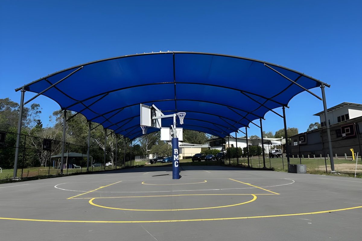 Rivermount College – Ground-Level View of Multi-Sport Court Under Blue Curved Tensile Canopy Ground-level view from one end of the sports court at Rivermount College, showing the blue curved tensile canopy overhead, the RMC branded court post with basketball hoop, yellow court lines, and surrounding suburban landscape