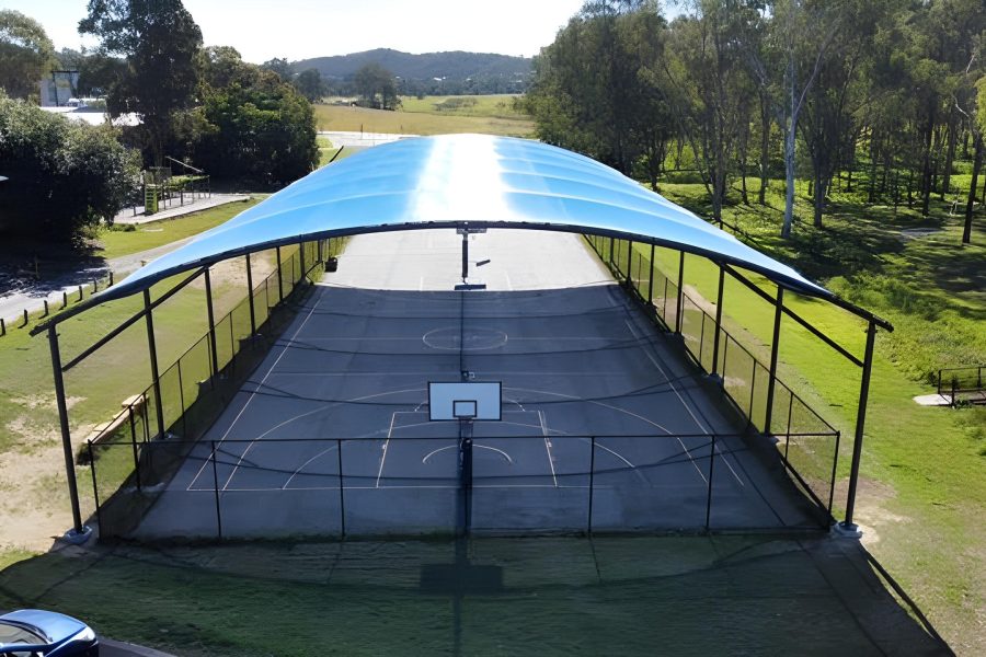 Rivermount College – Aerial End View of Blue Curved Tensile Canopy Over Multi-Sport Court Aerial end-on view of a blue curved tensile shade canopy covering a multi-sport court at Rivermount College, showing the full length of the court with basketball hoop visible underneath, surrounded by rural landscape and trees
